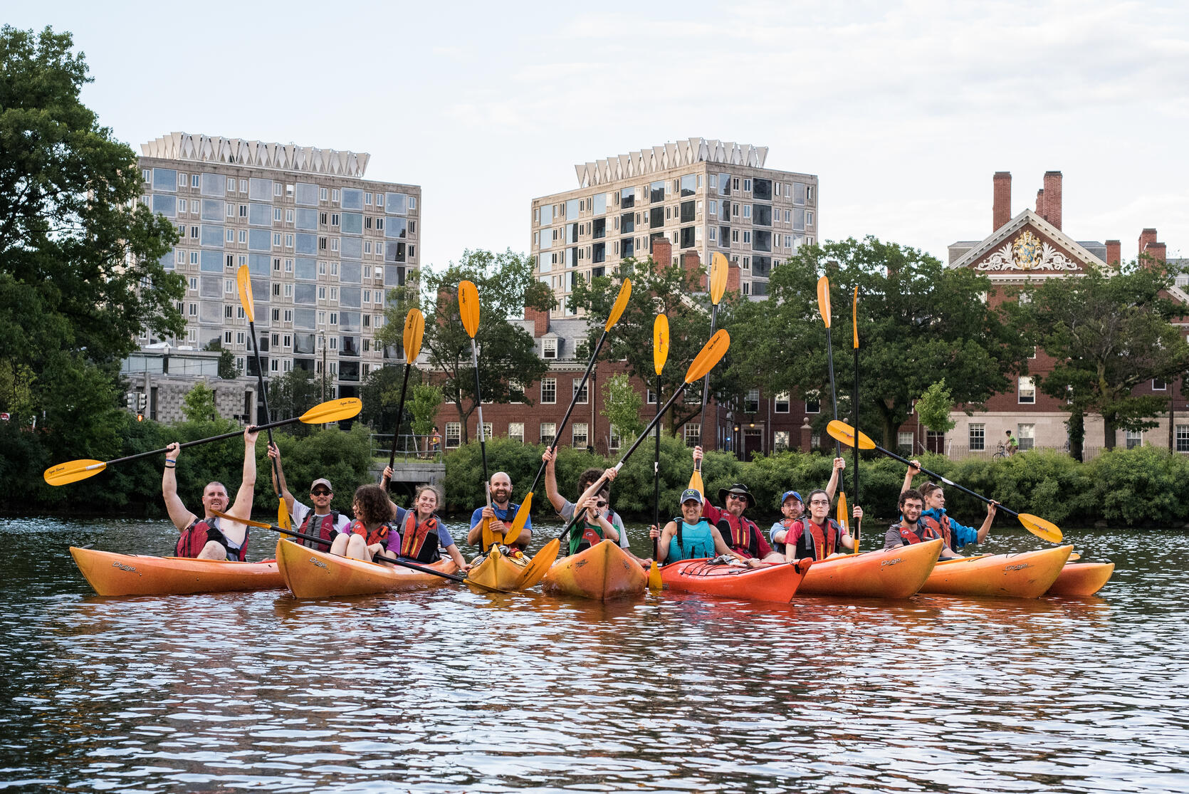 A group of kayakers on a river smile for the camera and raise their paddles.