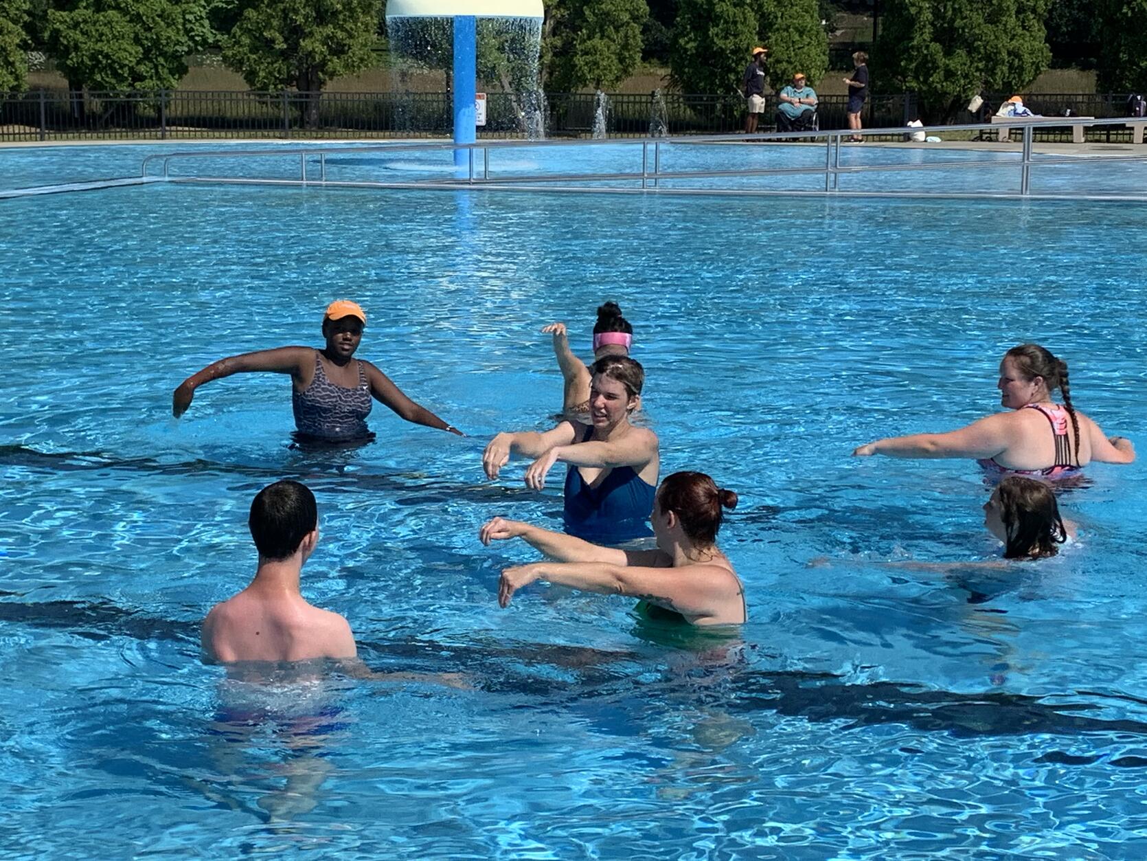 A group of people are standing in a circle around an aquatics instructor in a pool. 