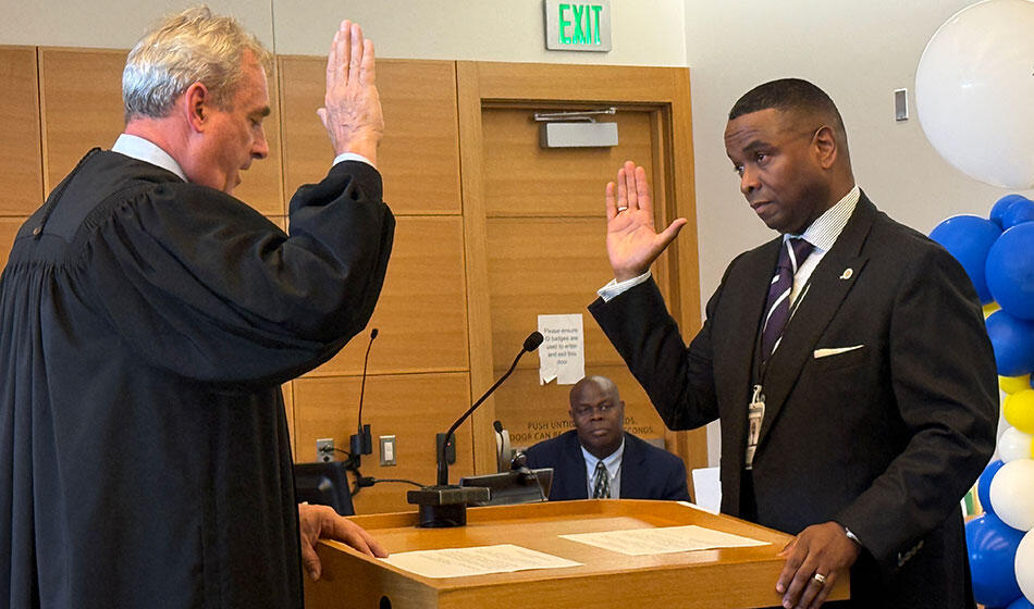 Two men participate in a swearing-in ceremony in a courtroom with an observer in the background.