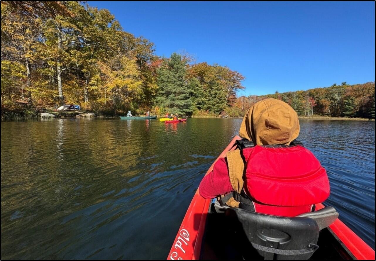 A person seated in the front of a kayak observes a group of paddlers on a tree-lined lake in autumn.