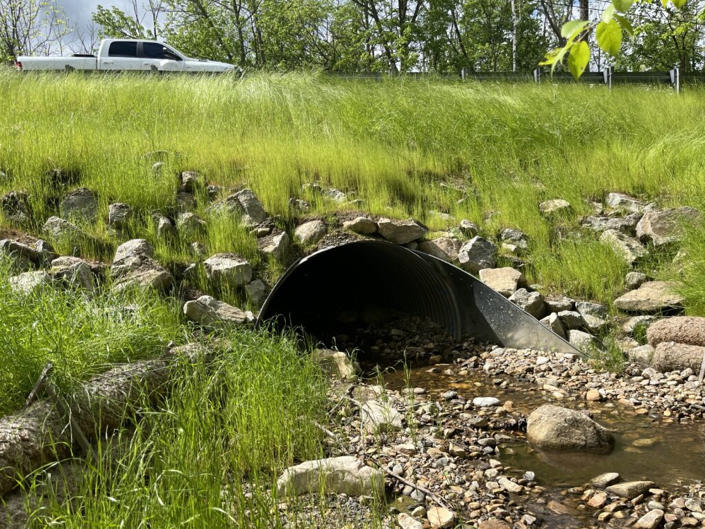 Water flowing through a culvert surrounded by green grass and a truck driving over it.