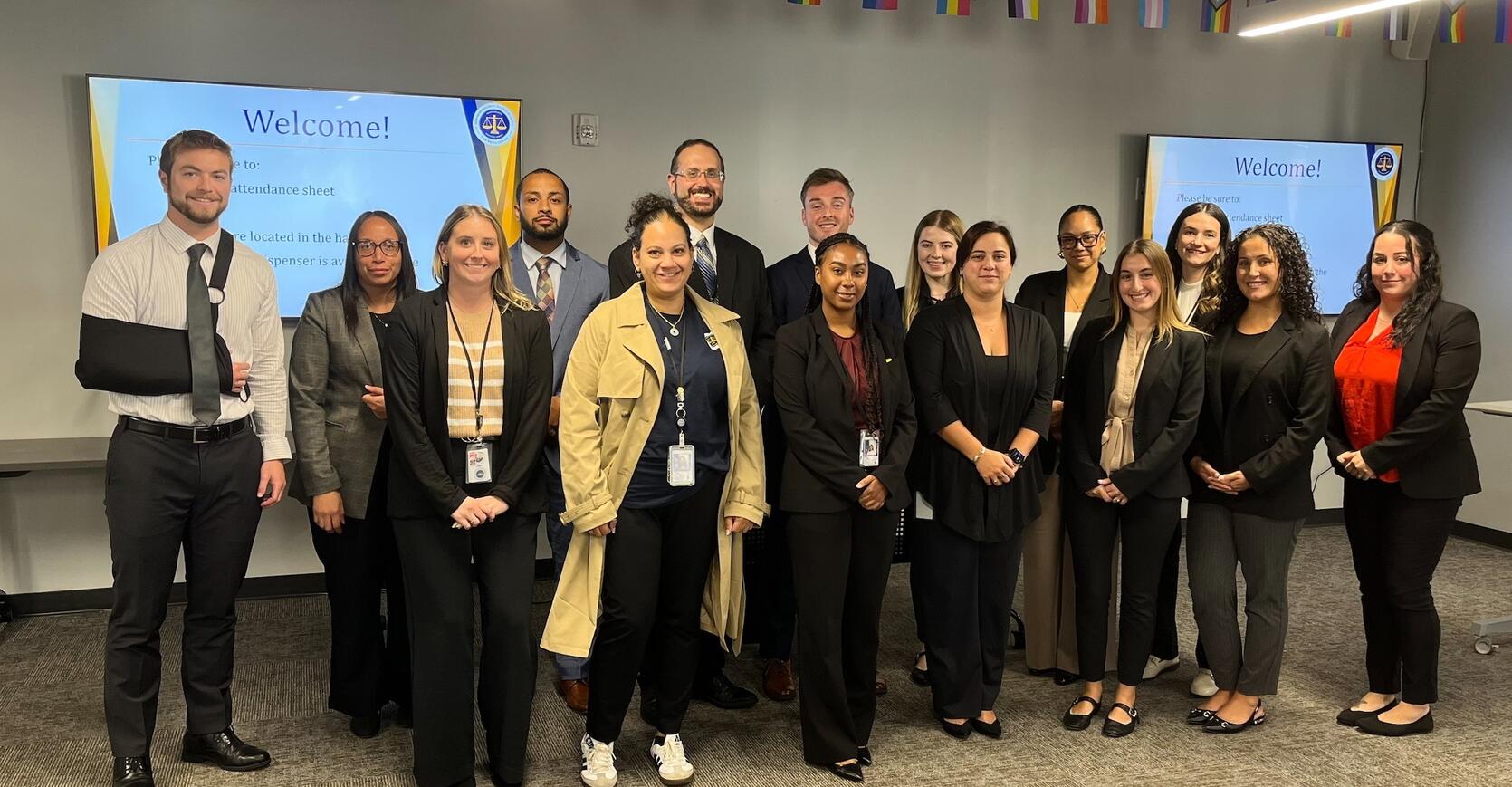 A group of 14 people in professional attire standing in a conference room with "Welcome!" displayed on screens in the background.