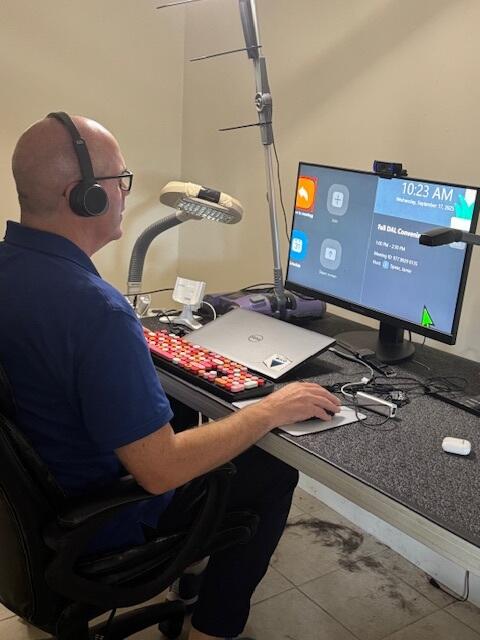 Side view of Erich working at his desk. He is a middle-aged white man, wearing glasses and headphones, and he’s viewing the details of an upcoming Zoom meeting in a magnified view on his large-screen display.