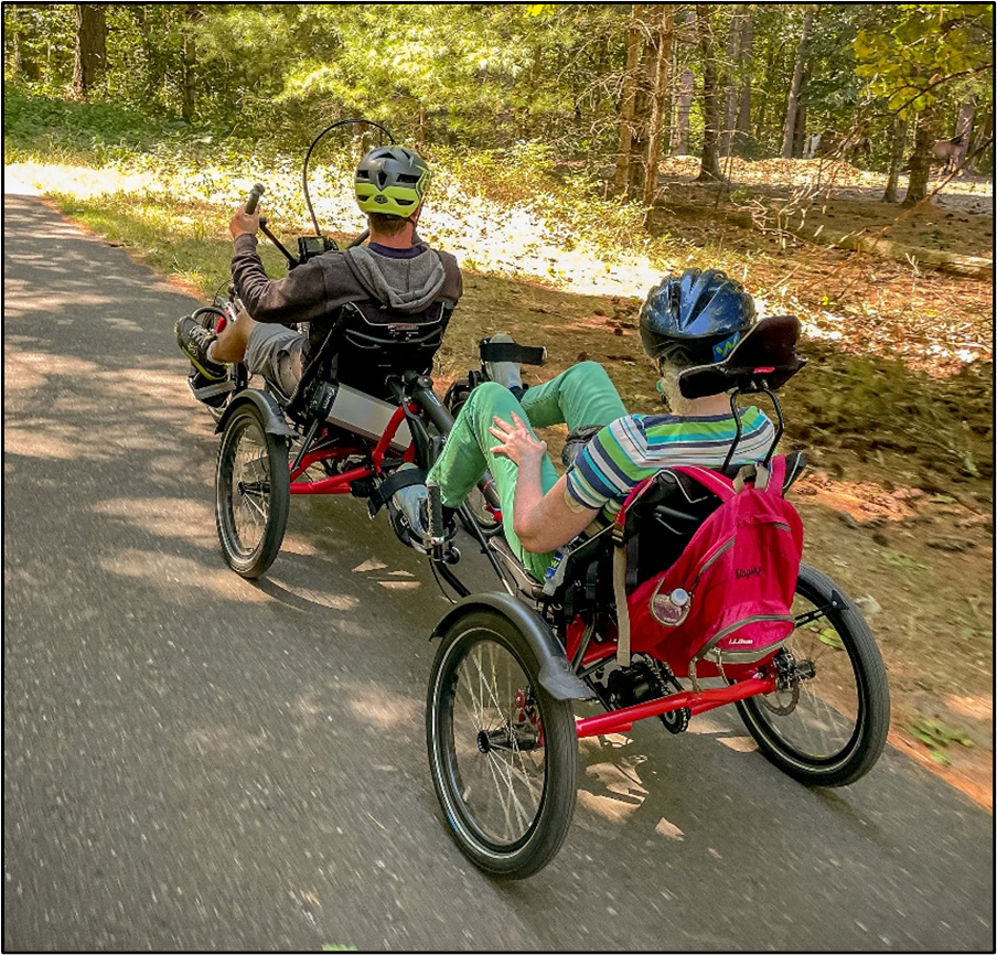 Two people riding a recumbent tandem cycle on a paved bike path are looking at the scenic woods.