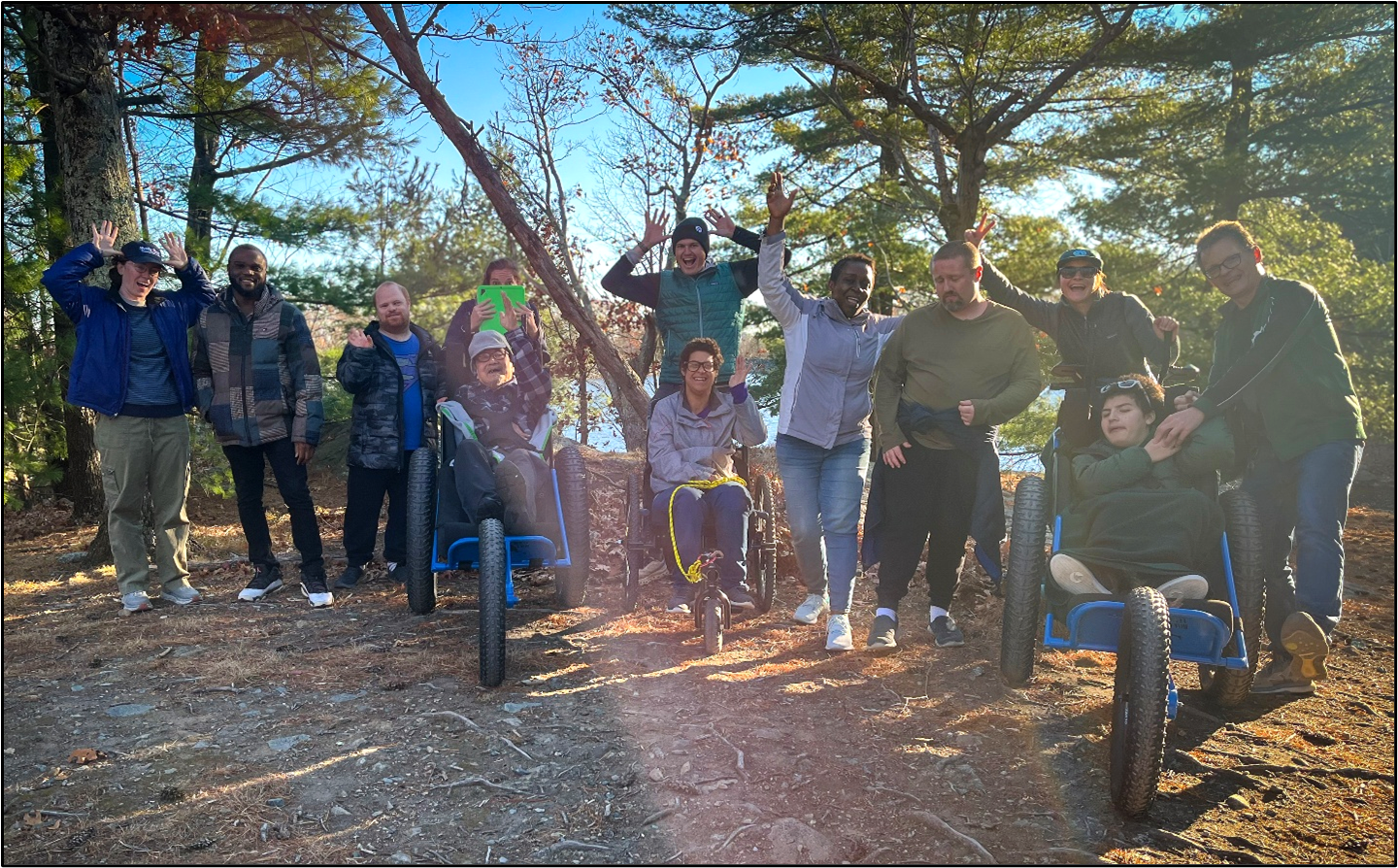 A group of hikers smile and wave on a dirt trail in the woods. Three are seated in all-terrain wheelchairs.