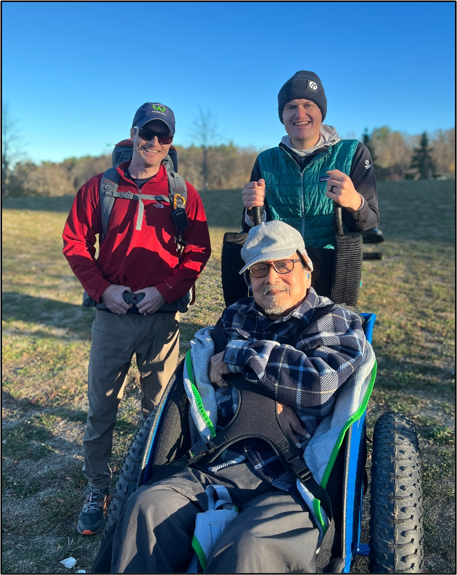 A hiker seated in an all terrain wheelchair and supporting staff smile on a dirt trail in the woods.