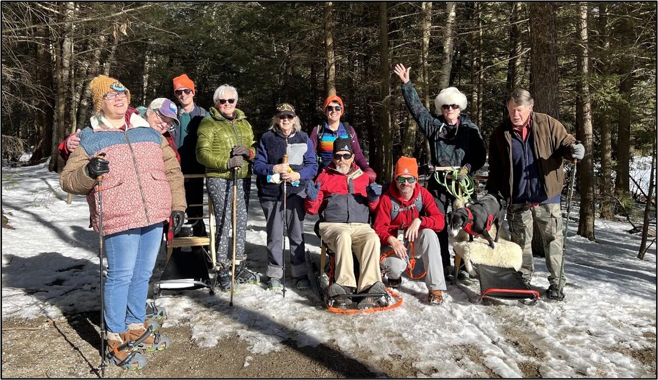 A group of hikers pose for a photo on a snowy trail in the woods. Some are using kick sleds and some are using walking sticks for support.  