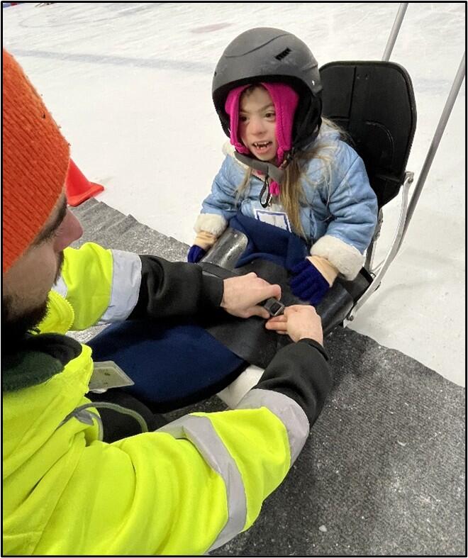 A young person seated on an ice sled smiles while UAP staff tie down a  lap blanket.