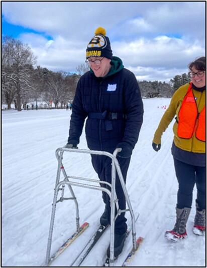 A person smiles while cross-country skiing. He is supported by a walker equipped with skis and program staff walking alongside.   