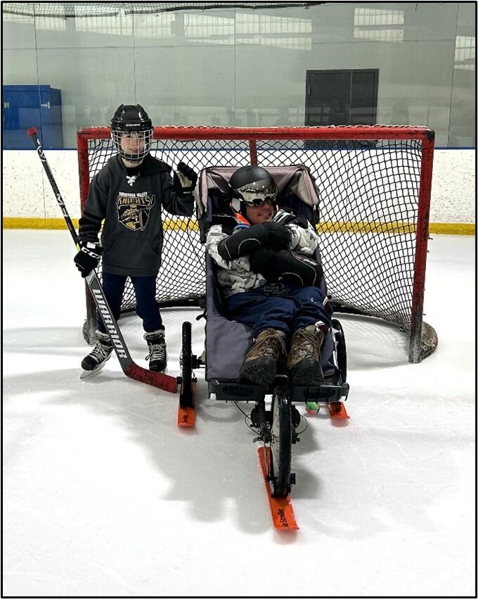 Two people in hockey gear pose for a photo in front on a hockey goal on the ice.  One person is seated in their adaptive push stroller with ski attachments. 