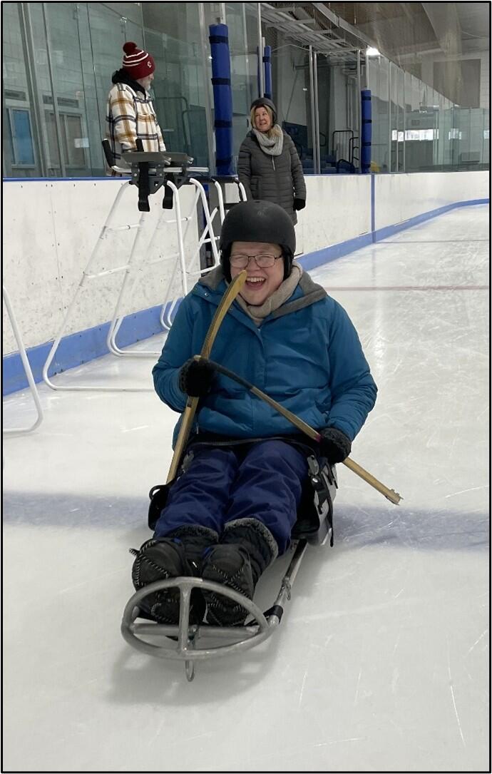 A person is laughing while propelling themselves on an ice sled with adaptive hockey sticks. 