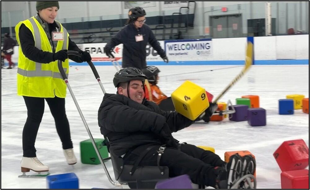 A person using an ice sled laughs while being pushed through foam blocks on the ice. He is trying to hit a block with a hockey stick.