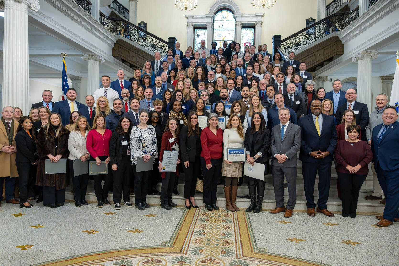 Large group photo of educators and school leaders from 63 Massachusetts Schools of Recognition standing on the Grand Staircase at the Massachusetts State House. Education Secretary Dr. Patrick Tutwiler and Commissioner Pedro Martinez stand with honorees holding certificates and legislators during the recognition ceremony.