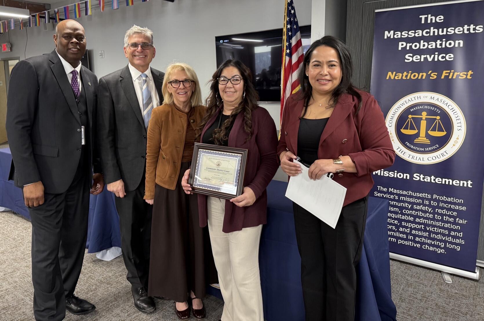 A group of 5 people pose with a woman in the middle holding a framed certificate