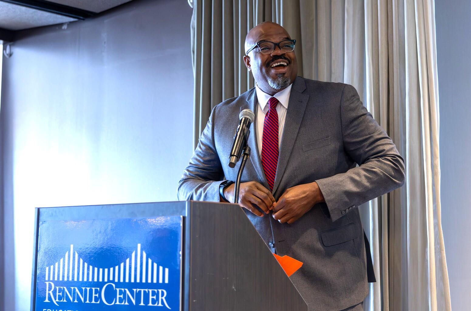 Secretary Tutwiler is wearing a suit with a red tie, standing at a podium with a sign that says Rennie Center