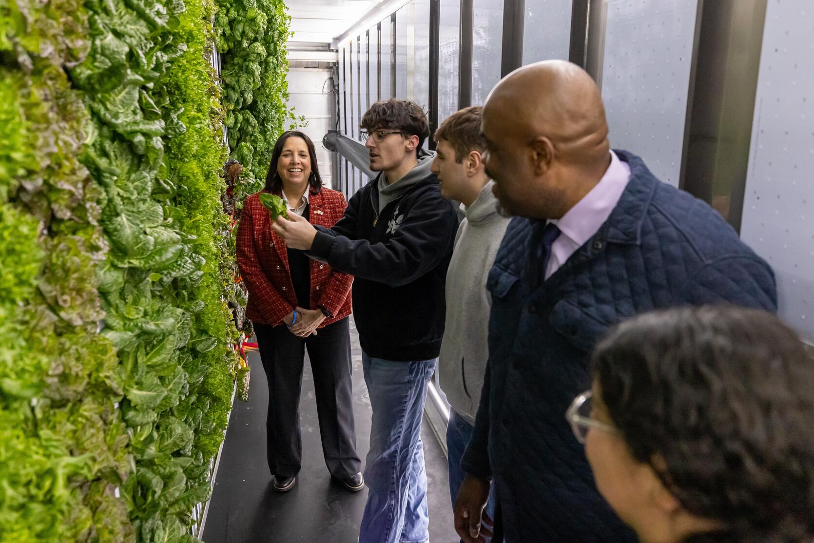Education Secretary Tutwiler and Lieutenant Governor Driscoll stand with a group of Watertown High School students inside a freight farm, in front of a large display of lettuce and kale growing on a vertical rack.