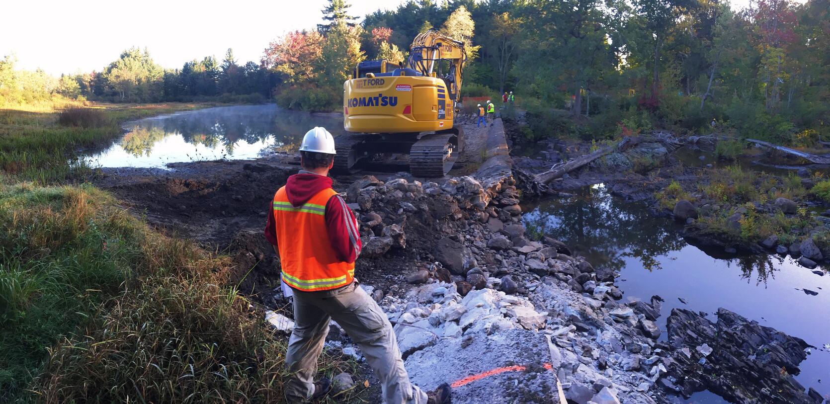 A person stands on the edge of a river with construction equipment in the background.