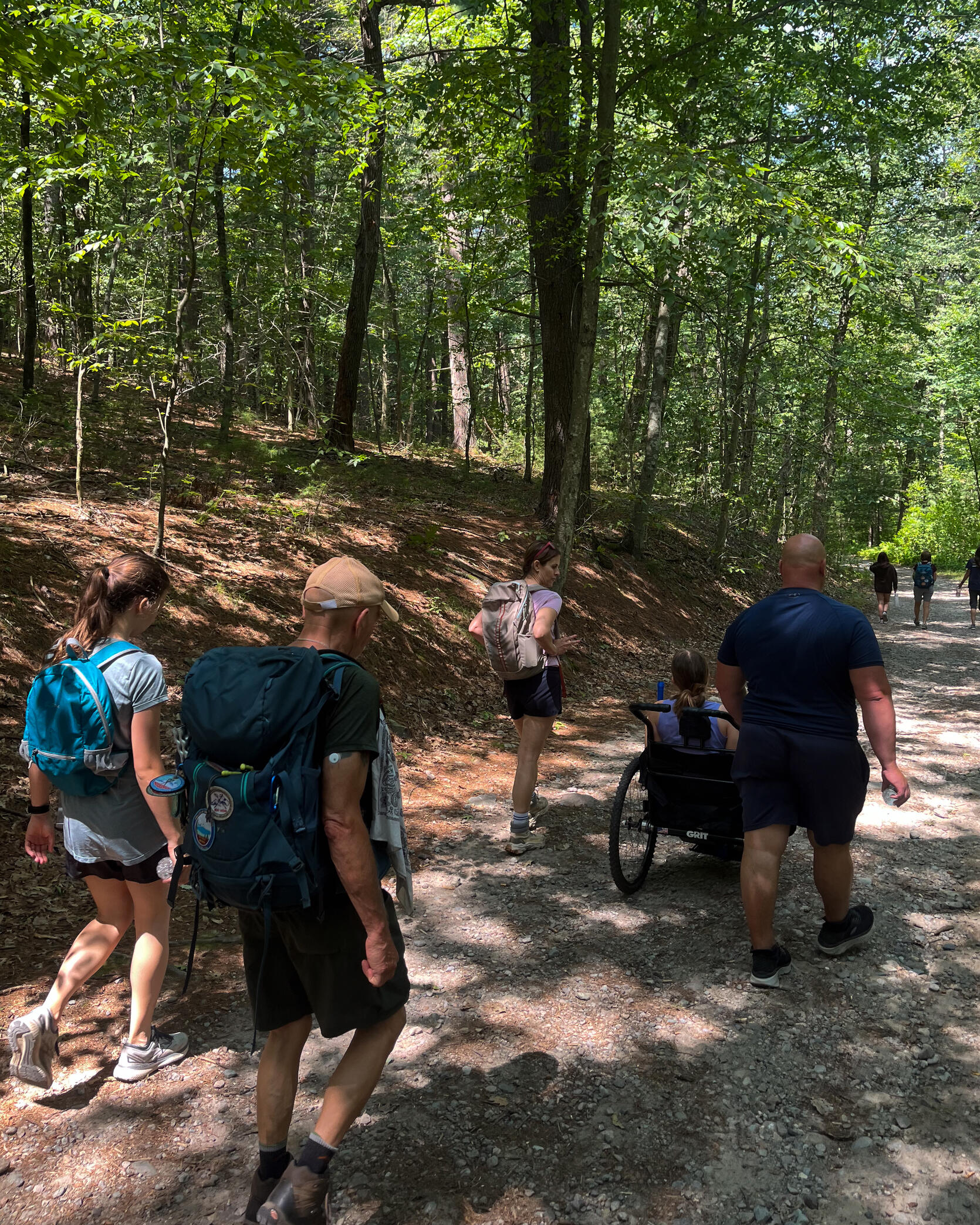 A group of people are hiking on a stone dust trail in the woods. One person is using an all terrain wheelchair. 