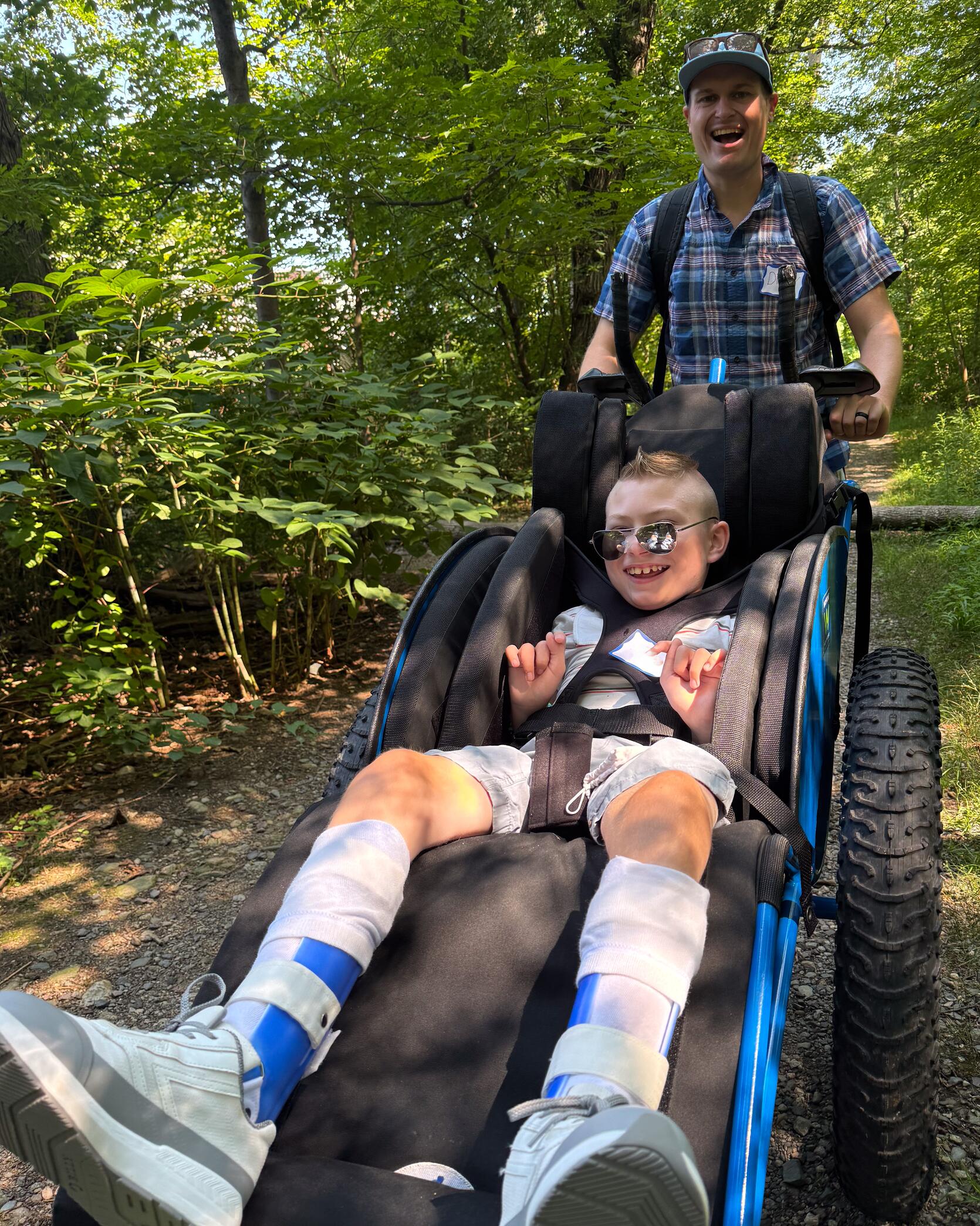 A boy smiles while being pushed on a stone dust trail through the woods in a Hoyt Racing Chair. 