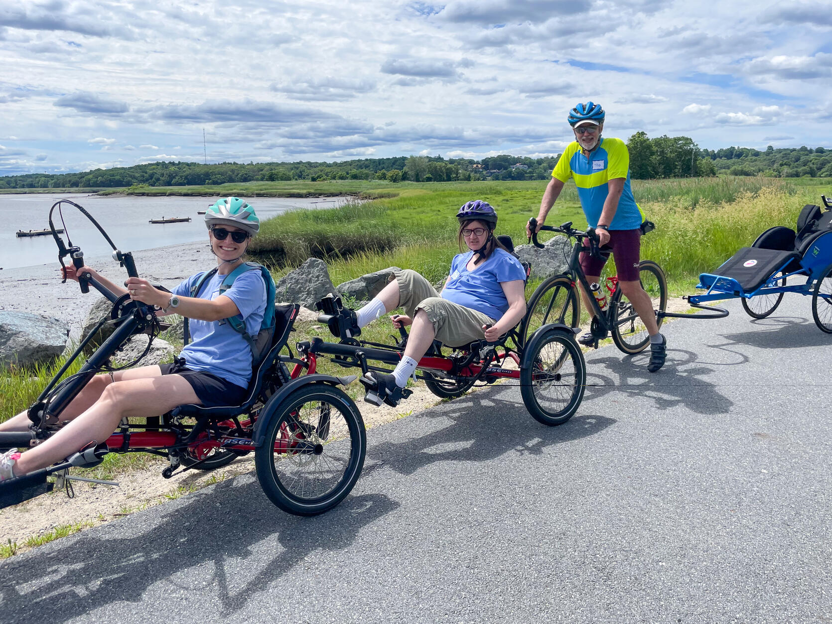 A group of people riding a variety of adaptive tandem bikes stop for a photo on a paved path along the water.  