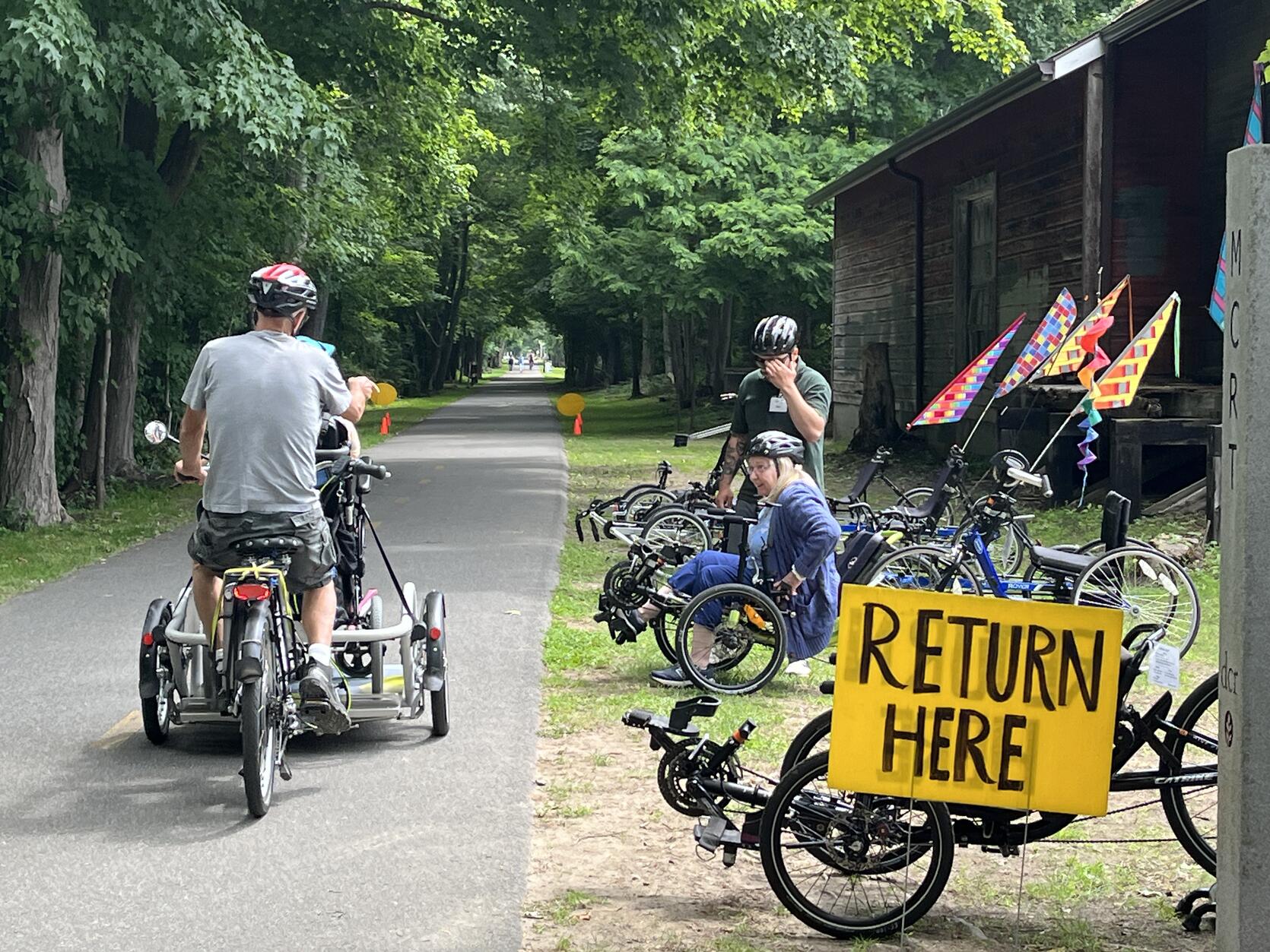 A wheelchair tandem cycle heads out on a tree-lined  paved bike path. A participant and staff sit on a recumbent bike among a line of adaptive cycles. 