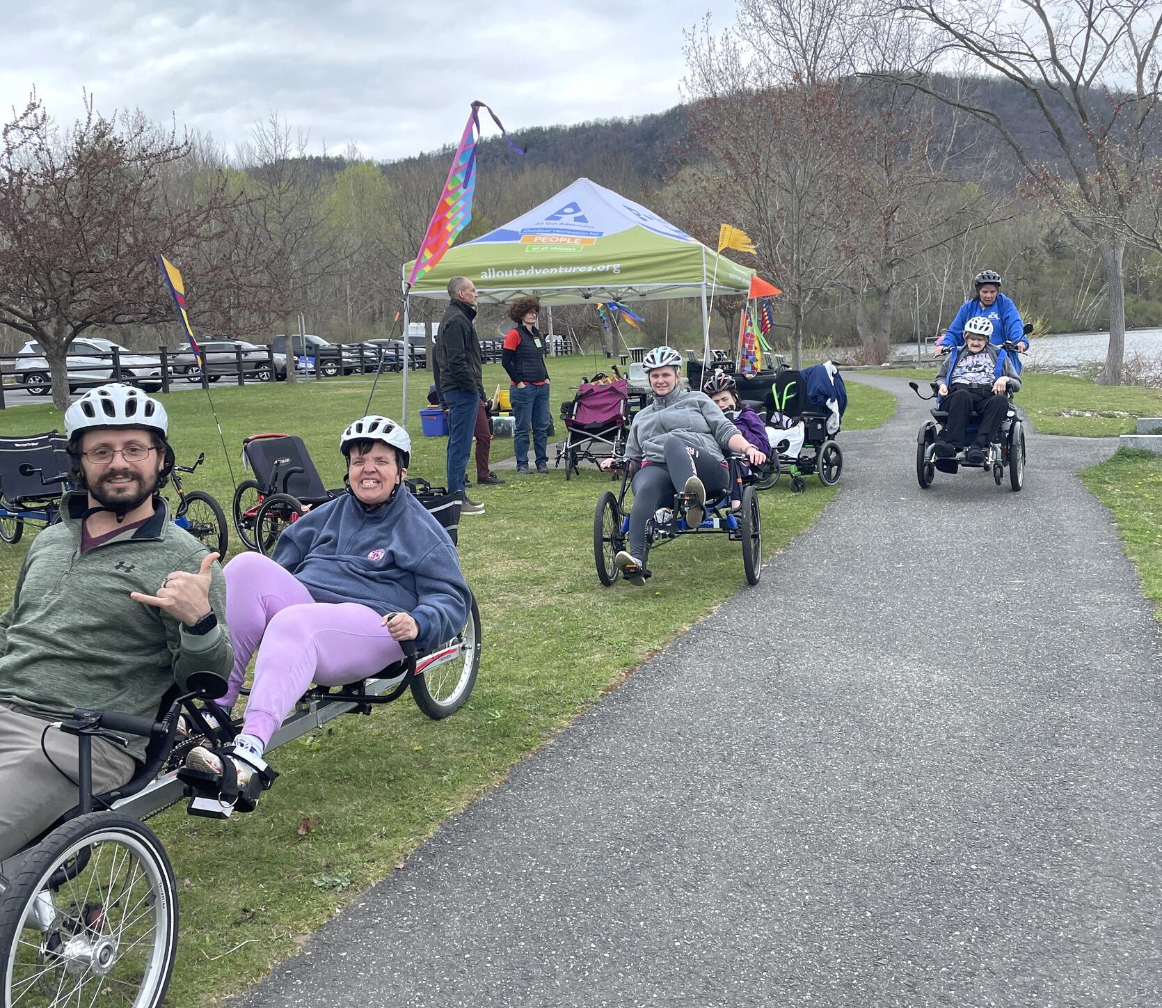 People using  a variety of adaptive cycles prepare to ride on a paved path next to a lake. 