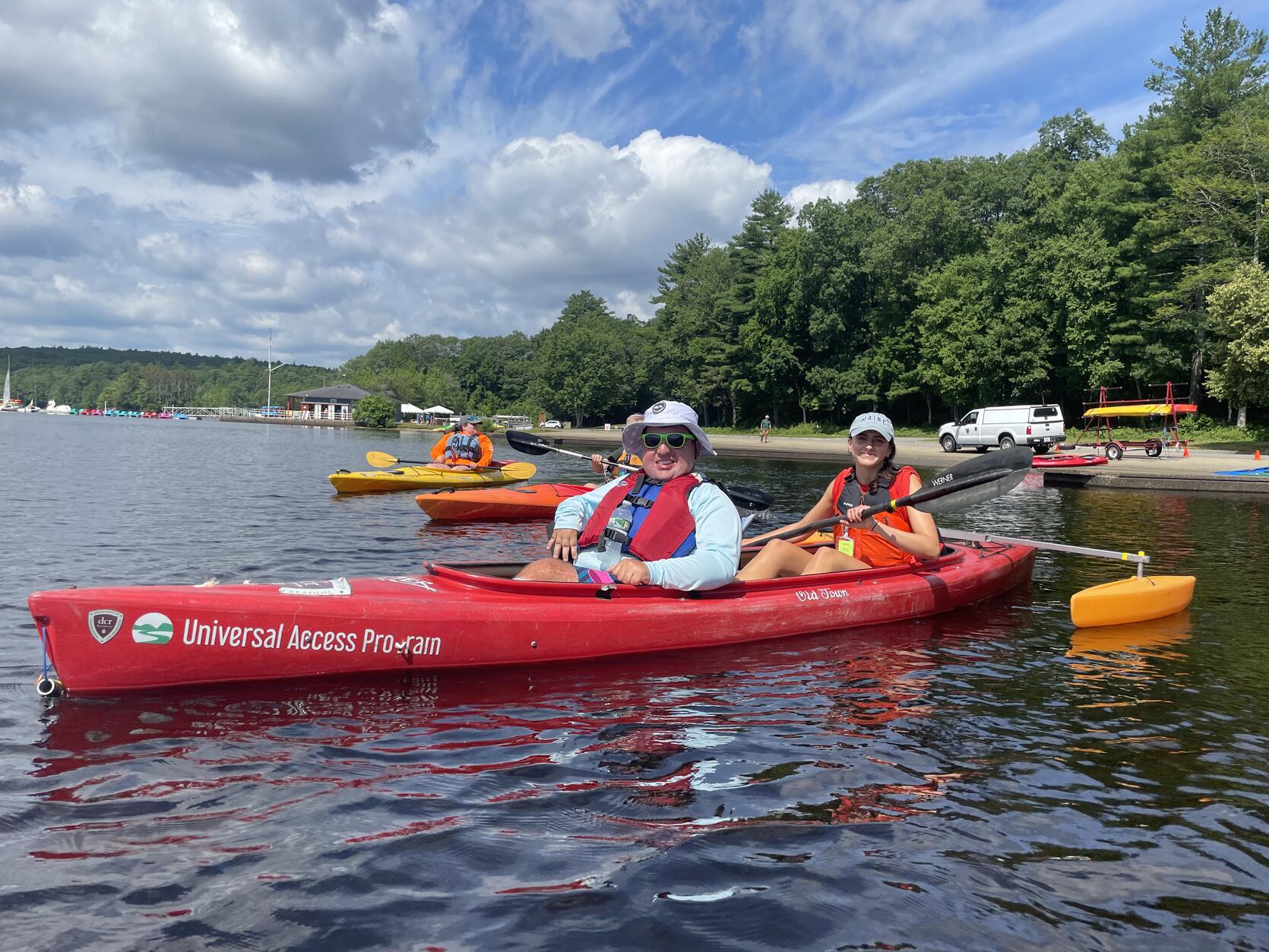 Two people smile while paddling in a tandem kayak equipped with pontoons on a lake. 