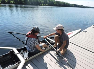 A person is seated in a double shell boat alongside a dock, is being assisted with oars.