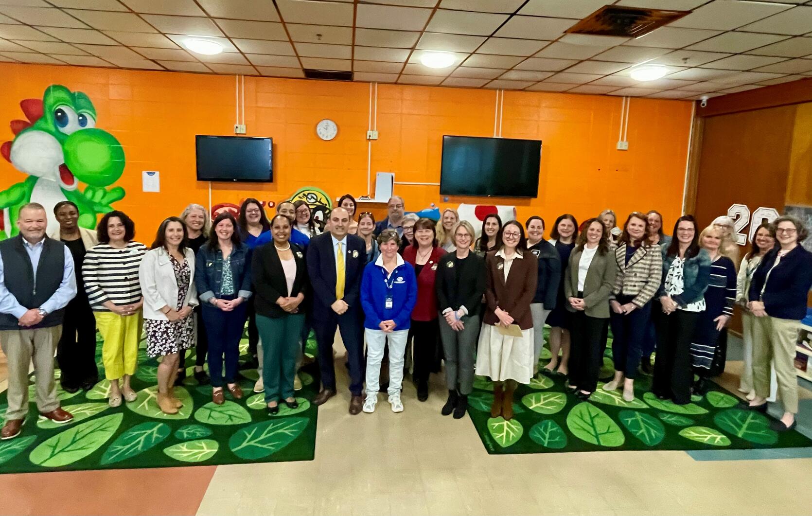 Education Secretary Steve Zrike stands with a group of local and state officials in an early child hood program in Pittsfield. Playful green children's rugs cover the tiled floor and a large cartoon Yoshi dragon is painted on the orange wall behind the group.
