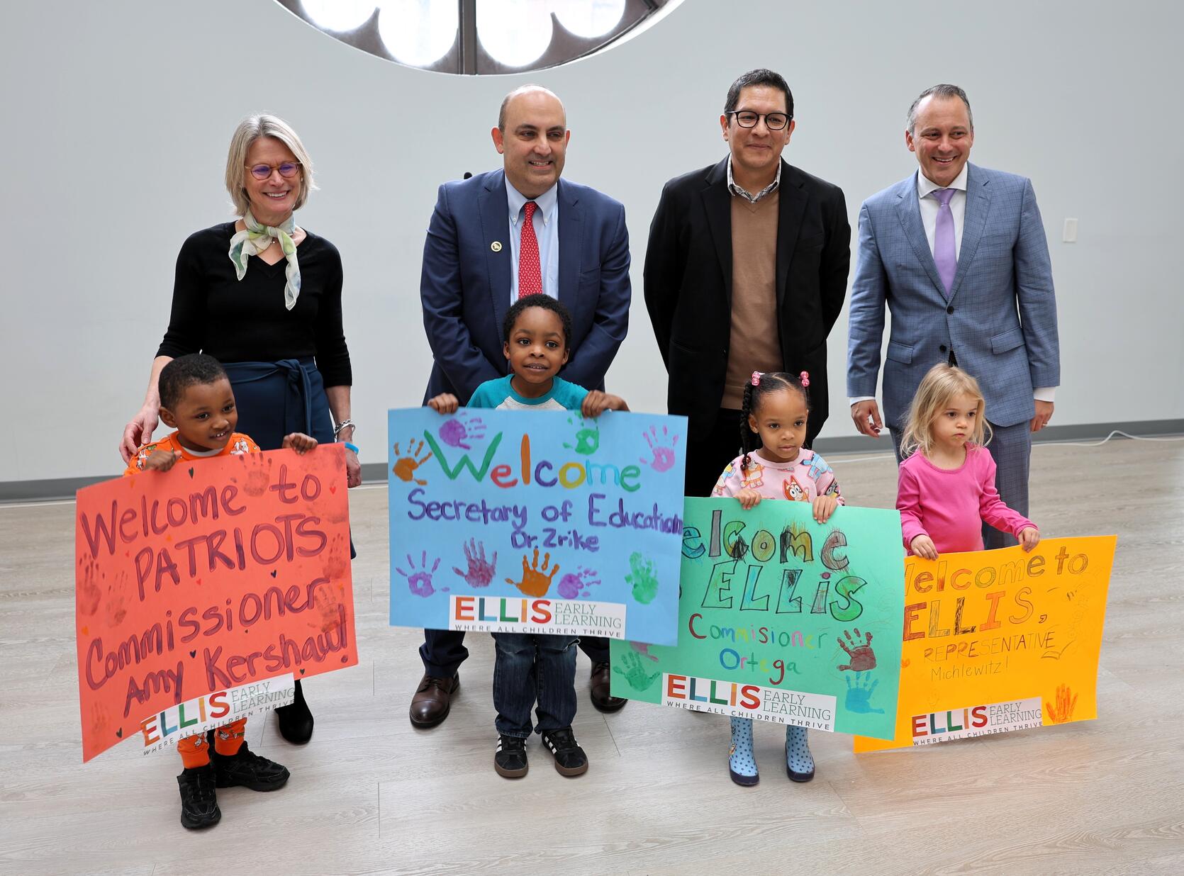 EEC Commissioner Kershaw, Secretary Zrike, DHE Commissioner Ortega and House Ways & Means Chair Michlewitz stand side by side in front of a while wall with four small children each holding brightly colored poster boards