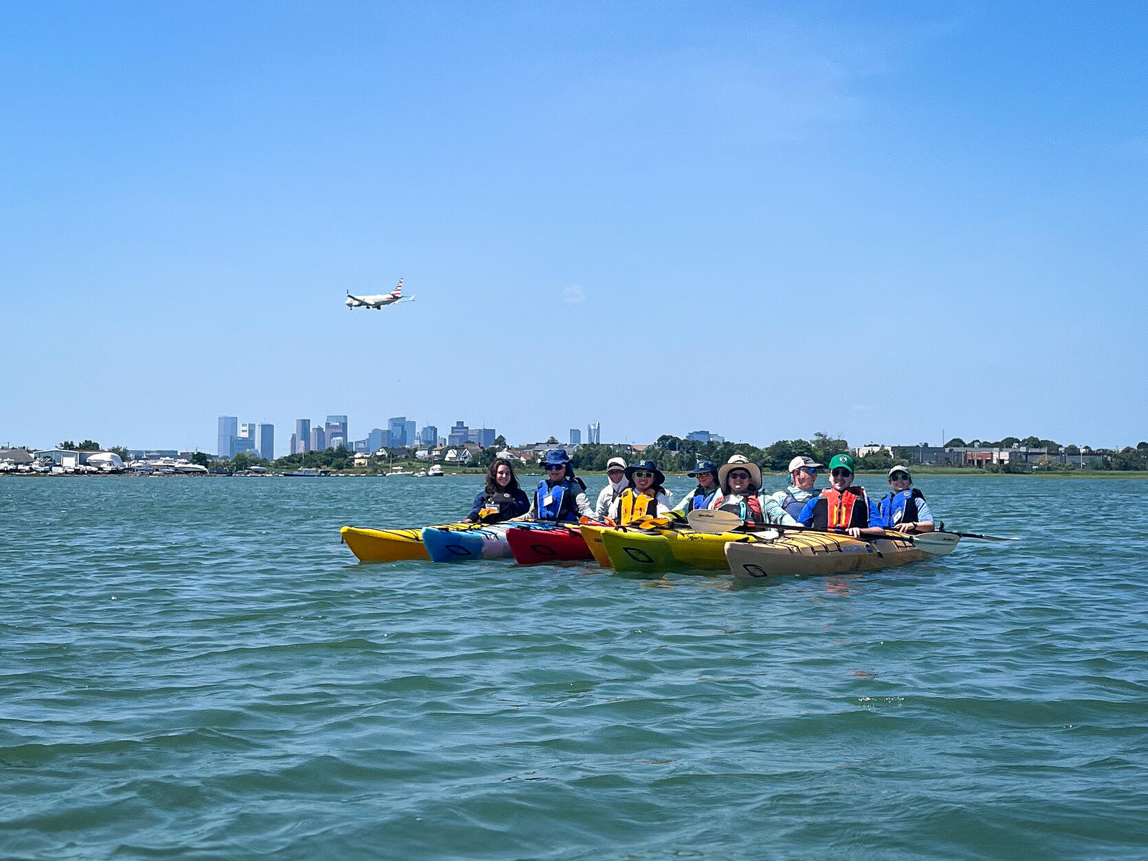 A group of kayakers in Boston Harbor pause for a photo as a plane flies over the Boston skyline. 