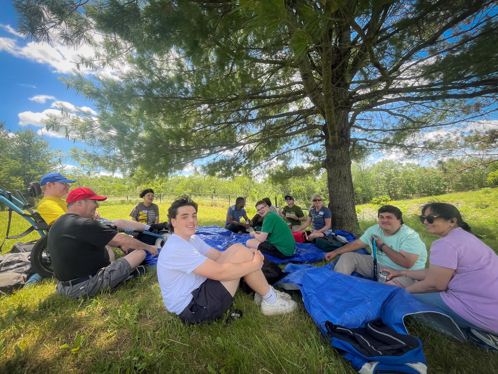 A group of hikers are resting under a tree. On person is seated in an all terrain wheelchair.
