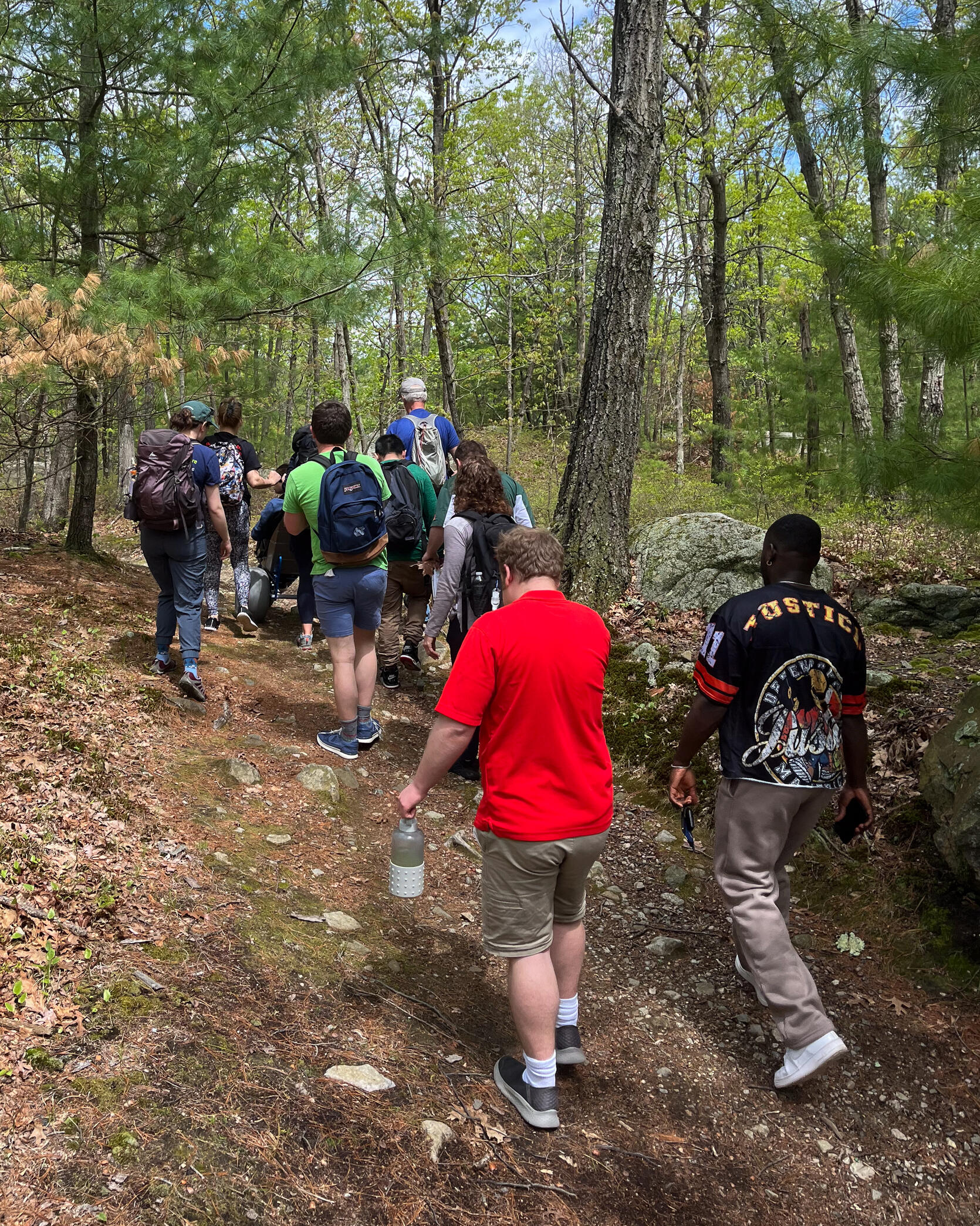 A group of people are hiking on trail in the woods. One person is using an all terrain wheelchair. 