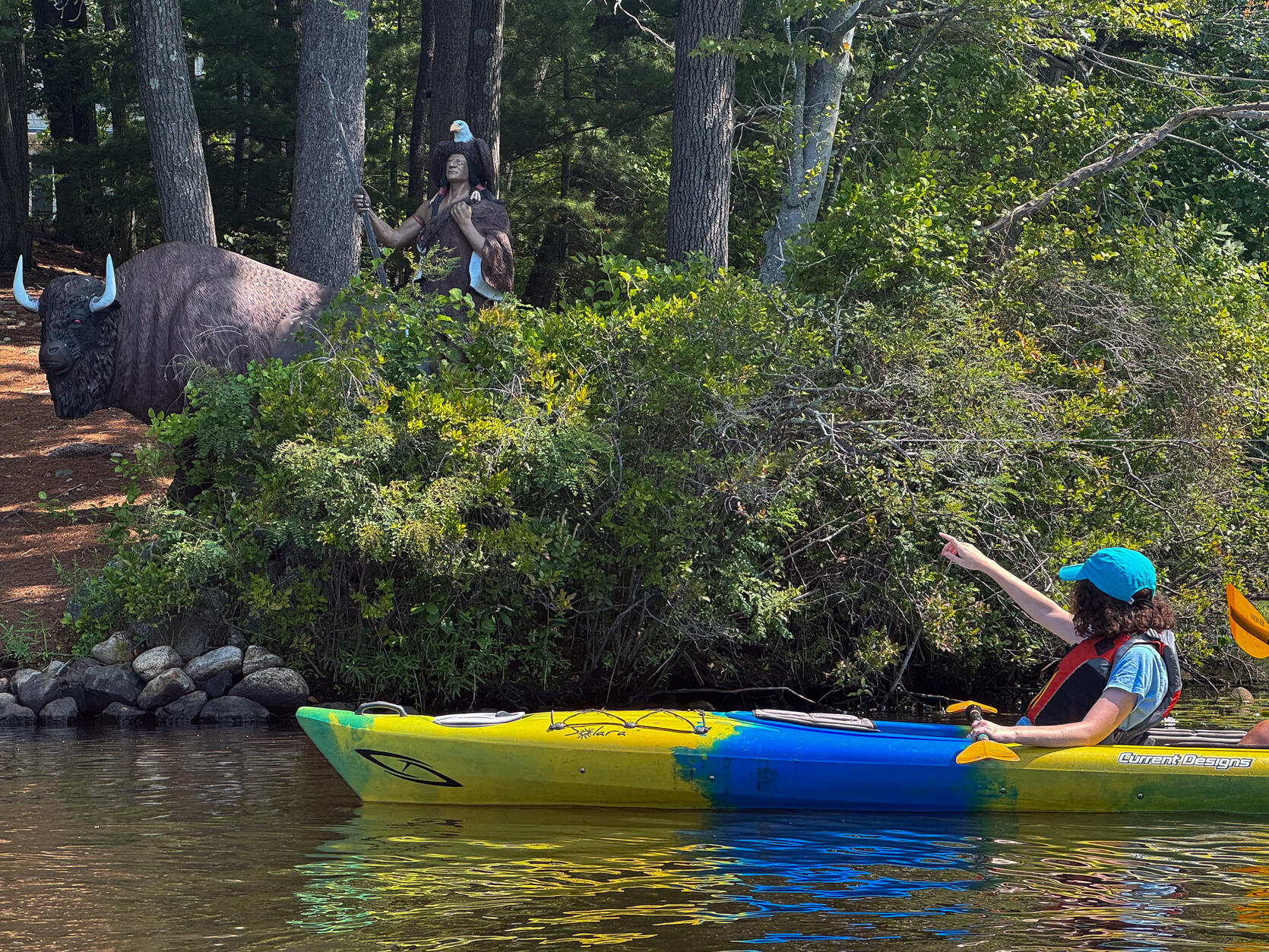 A kayaker pauses and points towards  the statute of a buffalo and Indian on the shore of a river.   
