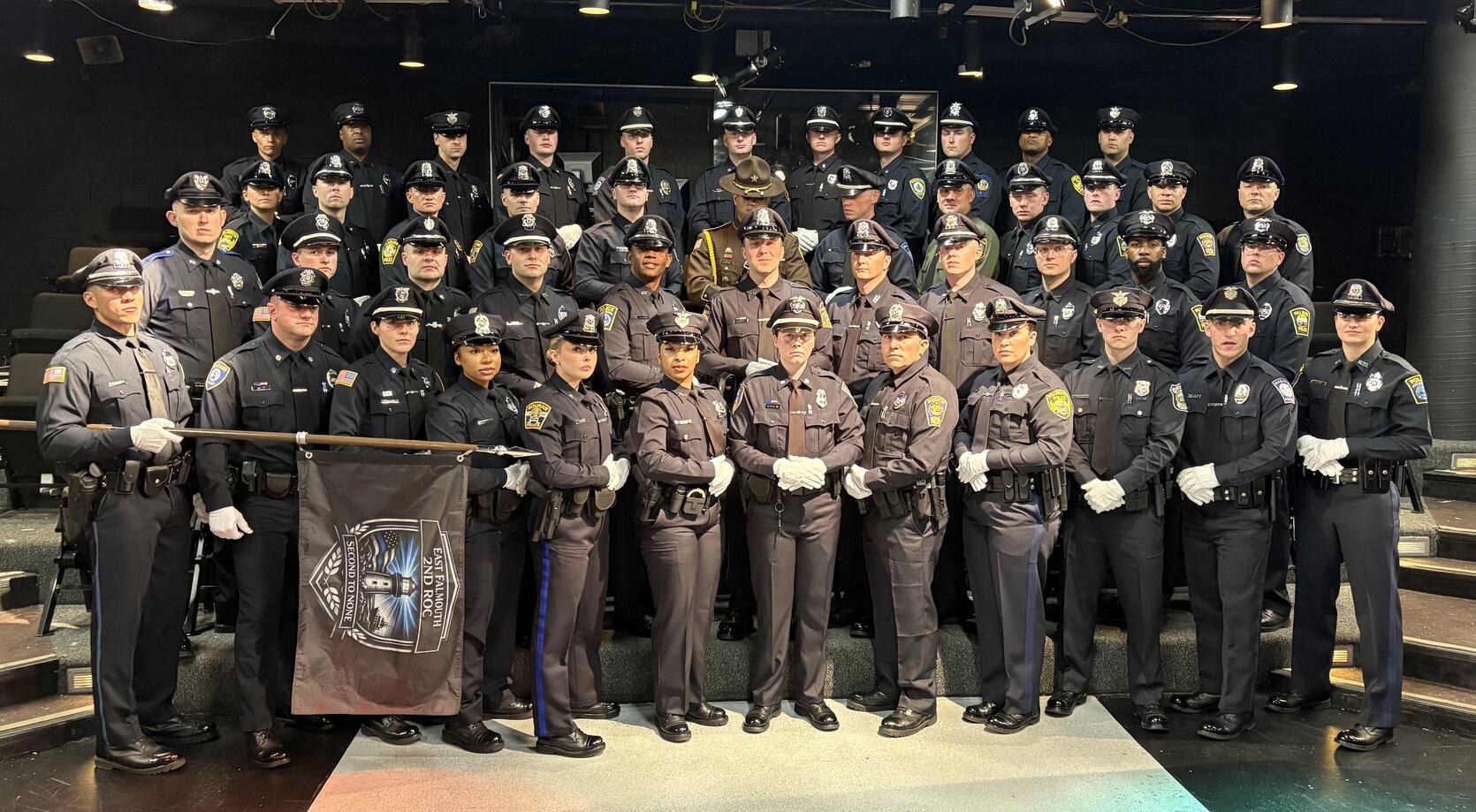 Large group of uniformed police officers posing indoors  for a graduation photo with a black flag displaying a police emblem in front.