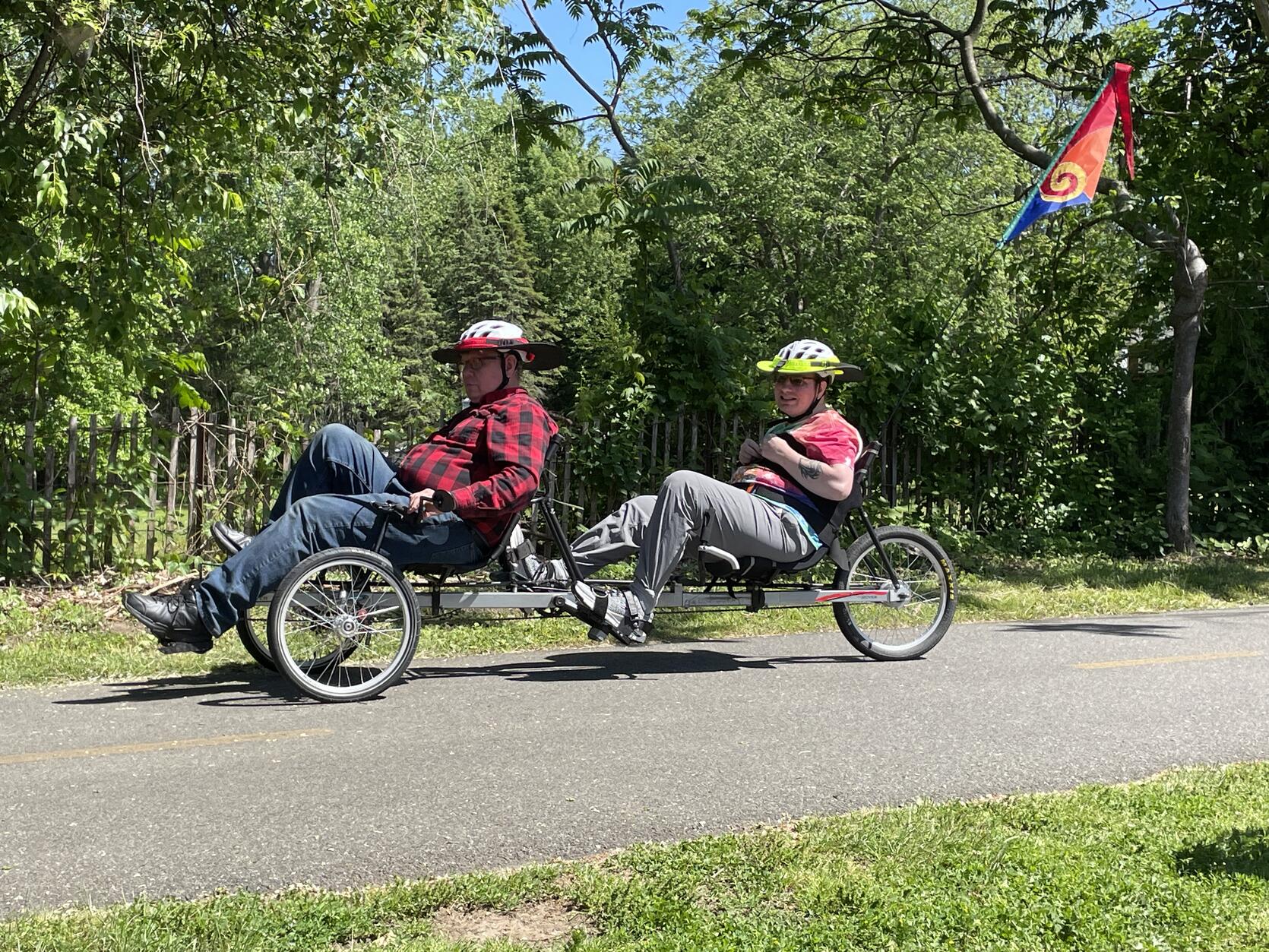 Two people a peddling on a  recumbent tandem cycle equipped with a bright flag, along a paved bike path. 