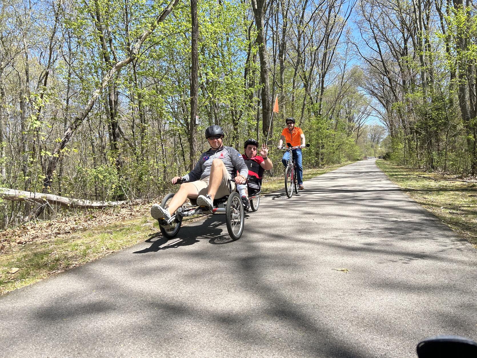 A person peddling on a recumbent tandem cycle waves while passing by on a paved bike trail.