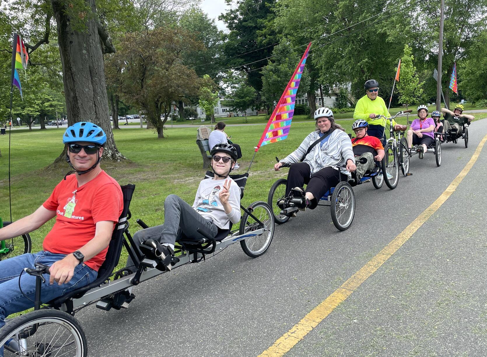 A line of people riding a variety of adaptive cycles on a paved bike trail stop and smile for a photo.