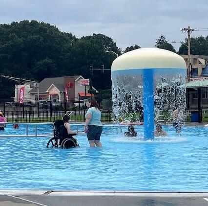 A group of people are playing in a pool equipped with an accessible ramp and splash pad. One person is using their wheelchair in the water. 