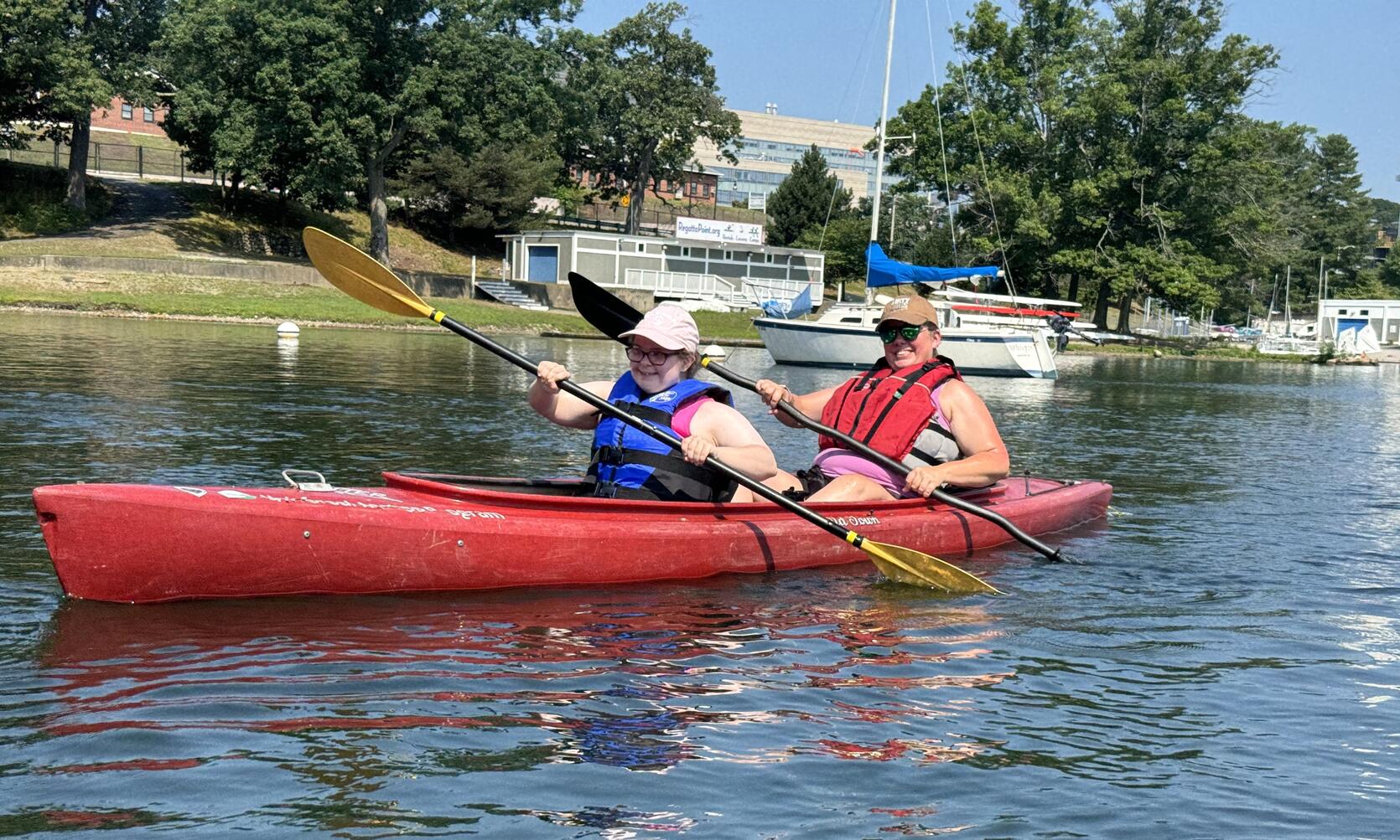 Two people in a tandem kayak smile while paddling past sailboats anchored on a lake shore. 