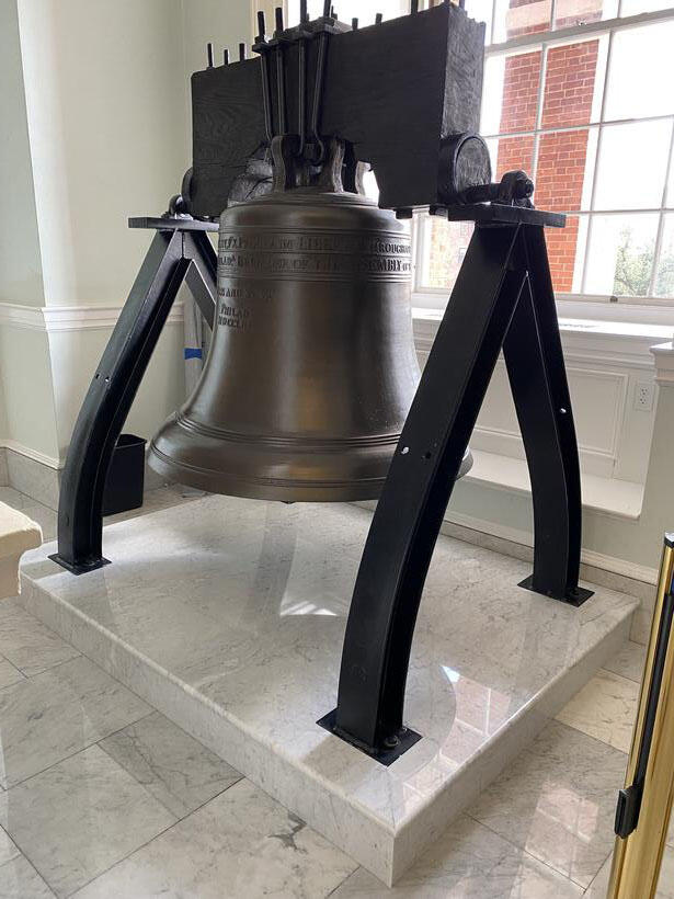 A large, bronze bell is displayed on a marble platform indoors, supported by sturdy black metal stands