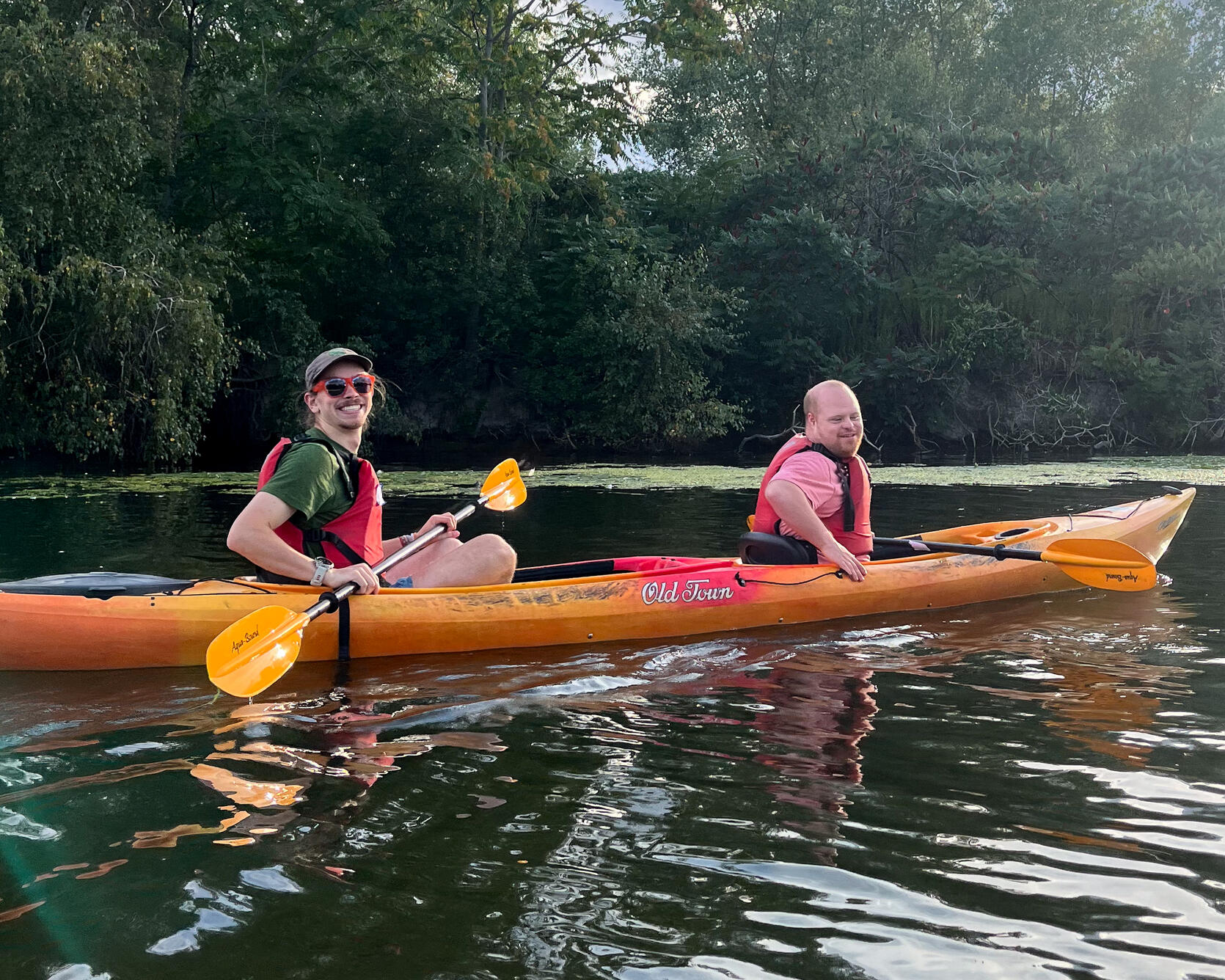 Two people paddling in a tandem kayak on a pond, smile for a photo. 