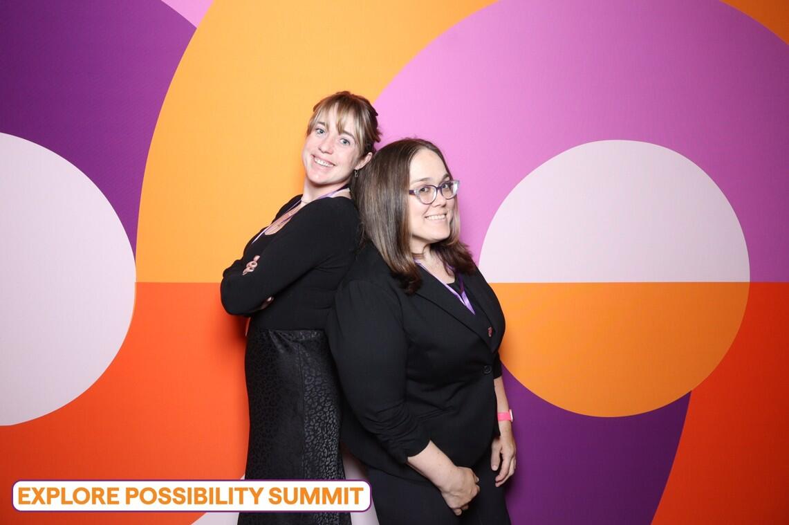 Two women standing back to back in front of a multicolored background. The woman on the right is Noelle. 