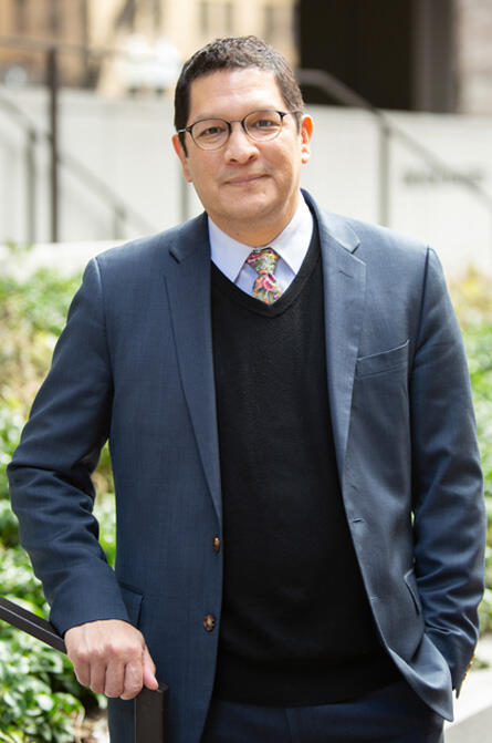 Higher Ed Commissioner Ortega wears a suit and poses for a photograph outdoors, leaning on a railing  and smiling
