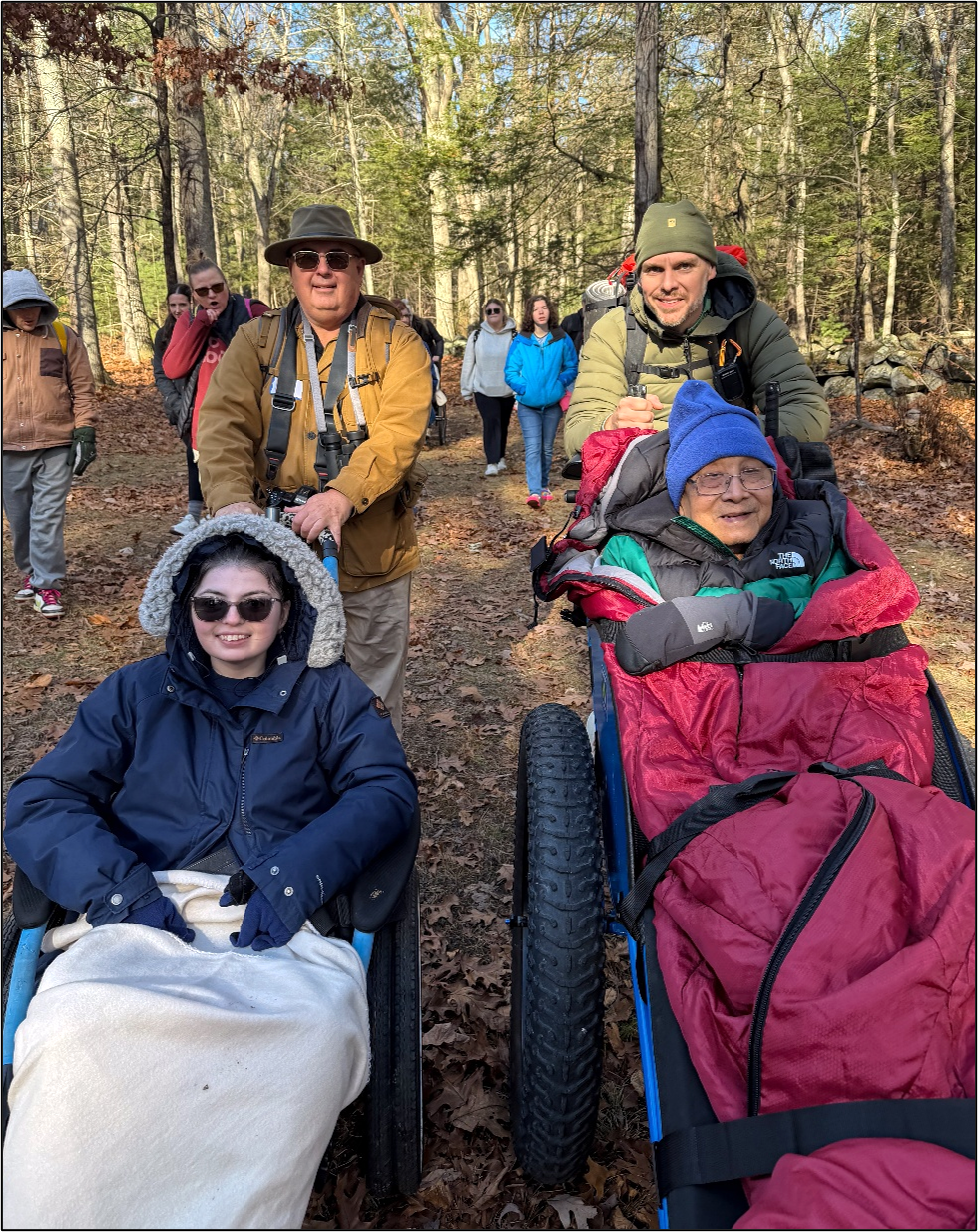 A group of people are hiking on a stone dust trail in the woods in autumn. Some are being pushed in in all-terrain wheelchairs.
