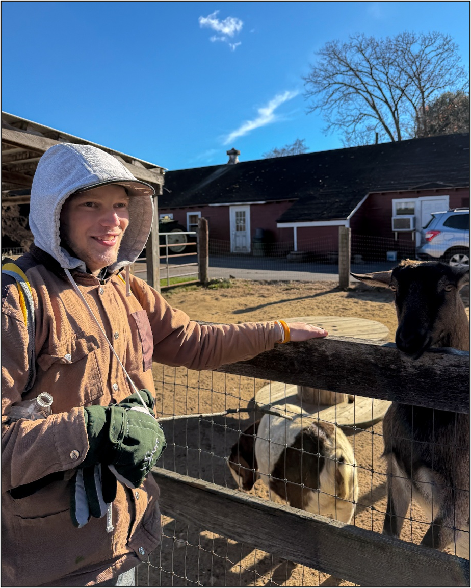 A young adult smiles while standing  next to a goat pen at a farm. 
