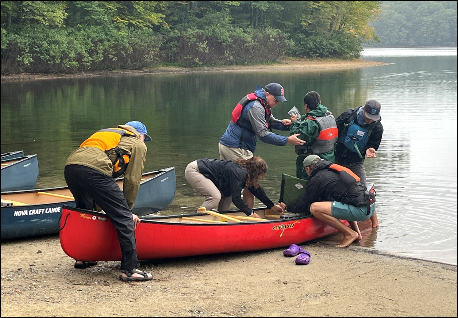 A group of people assist a participant into a canoe equipped with an adaptive seat on a lakeshore.