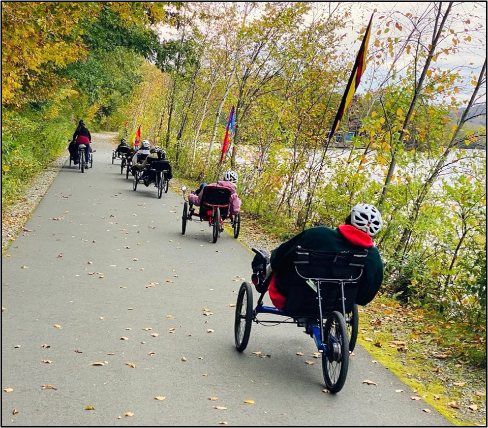 A group of people are riding recumbent trikes on a paved  trail along a lake in autumn.  