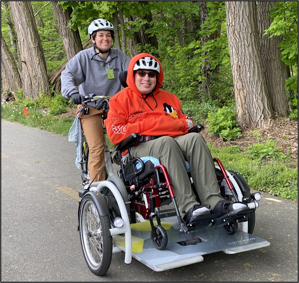 Two people are riding a wheelchair tandem cycle on a paved trail lined with trees.