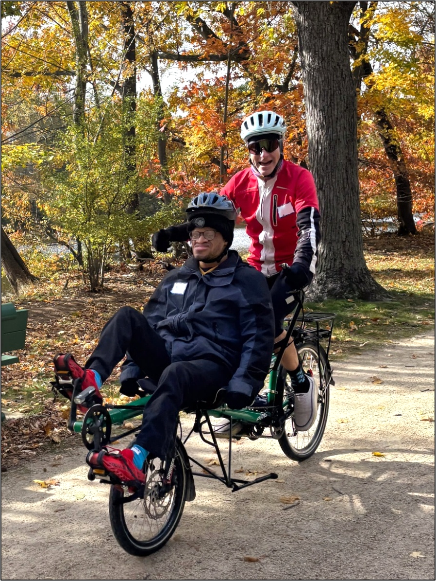 Two people are riding a semi-recumbent tandem cycle on a stone dust trail lined with autumn trees. 